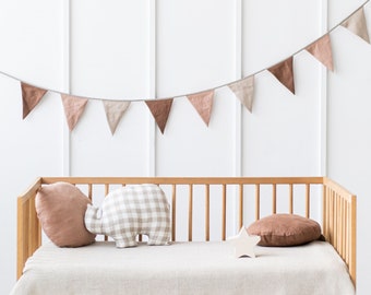 May include: A wooden crib with a white blanket and two pillows. One pillow is brown and the other is checkered. A string of brown and white flags hangs above the crib.