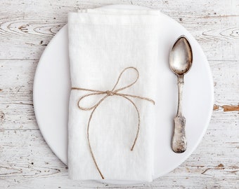 May include: A white linen napkin tied with twine, placed on a white plate with a silver spoon. The plate and napkin are on a white wooden surface.