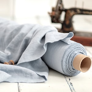 May include: A roll of light blue linen fabric with a soft, textured appearance. The fabric is partially unrolled, revealing its delicate weave. Two wooden spools of thread sit nearby, suggesting a sewing or crafting theme. An antique sewing machine is in the background.