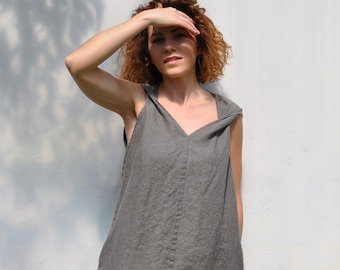 May include: A sleeveless, gray linen dress with a hood. The dress has a V-neck and falls to the knees. The person is standing in front of a light gray wall.