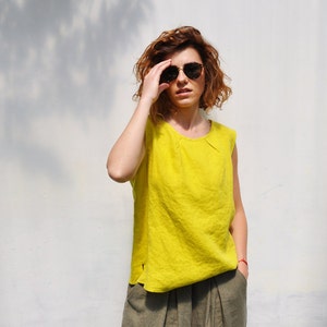 May include: A woman wearing a bright yellow sleeveless top and olive green linen trousers. She is wearing sunglasses and has her hair pulled back.