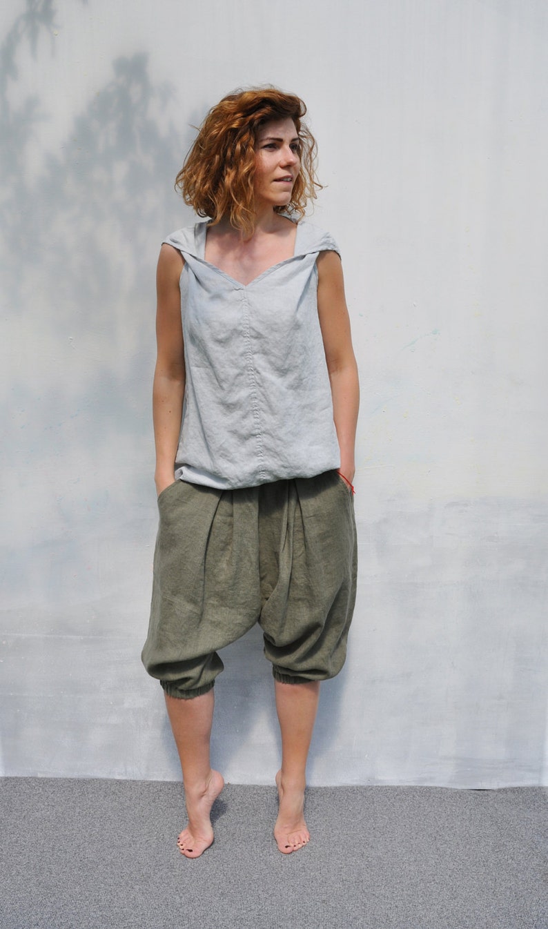 May include: A woman wearing a light gray linen sleeveless top with a hood and olive green linen pants. The pants have a loose, baggy fit and are gathered at the ankles.