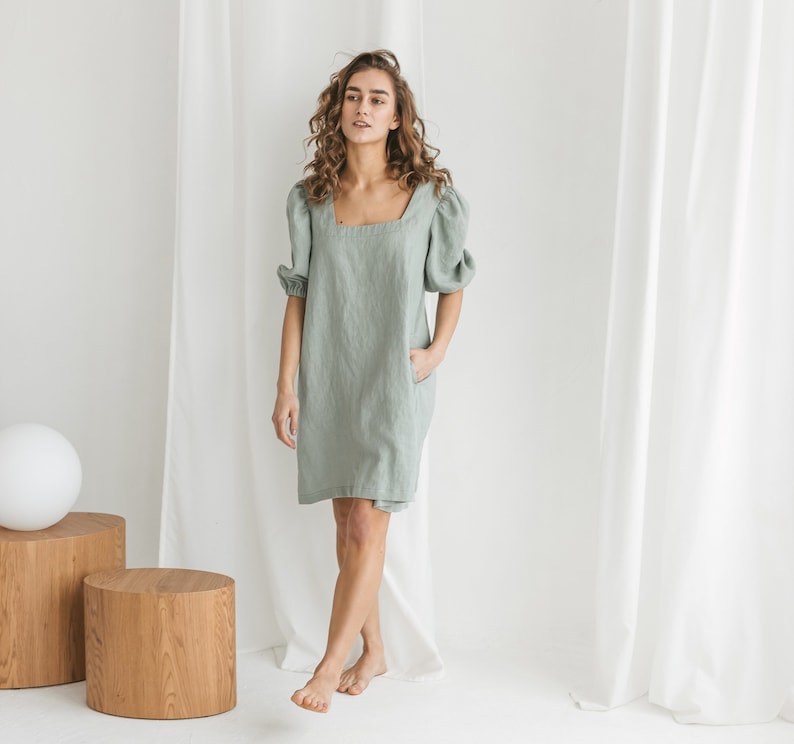 May include: A woman wearing a sage green linen dress with puffed sleeves. The dress has a square neckline and a relaxed fit. The woman is standing in front of a white backdrop.