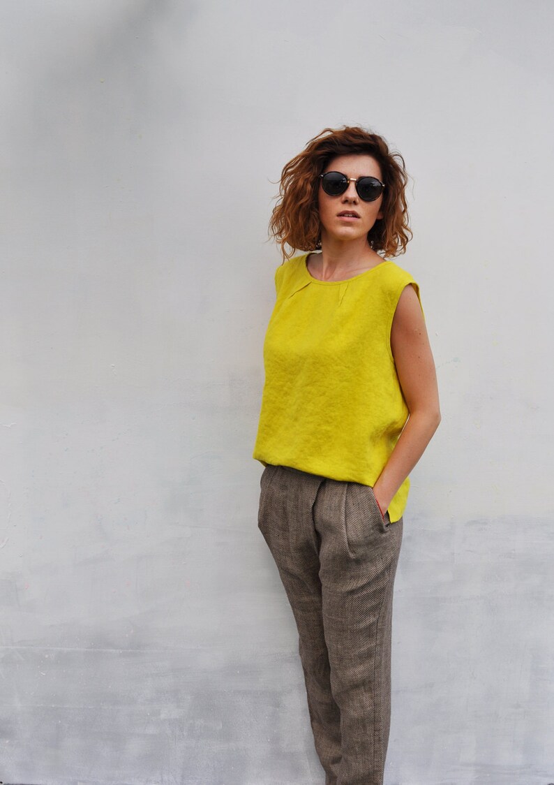 May include: A woman wearing a bright yellow sleeveless top and brown trousers. She is wearing round sunglasses and has curly brown hair.
