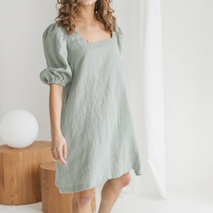 May include: A sage green linen dress with a square neckline and puffed sleeves. The dress has a relaxed fit and falls to the mid-thigh.