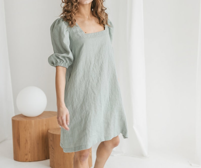 May include: A sage green linen dress with a square neckline and puffed sleeves. The dress has a relaxed fit and falls to the mid-thigh.