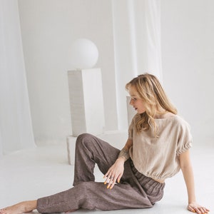 May include: A woman wearing a light brown linen top with a gathered elastic waistband and brown striped linen pants sits on a white floor.