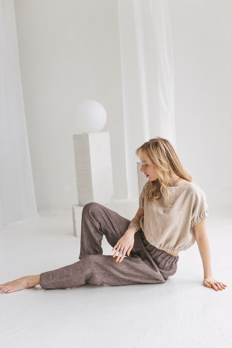 May include: A woman wearing a light brown linen top with a gathered elastic waistband and brown striped linen pants sits on a white floor.