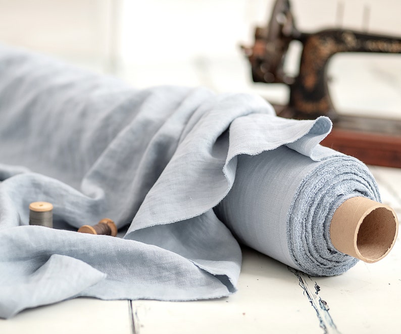 May include: A roll of light blue linen fabric with a soft, textured appearance. The fabric is partially unrolled, revealing its delicate weave. Two wooden spools of thread sit nearby, suggesting a sewing or crafting theme. An antique sewing machine is in the background.