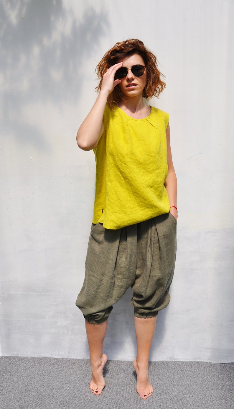 May include: A woman wearing a bright yellow sleeveless top and olive green linen trousers. She is wearing sunglasses and has her hair pulled back.