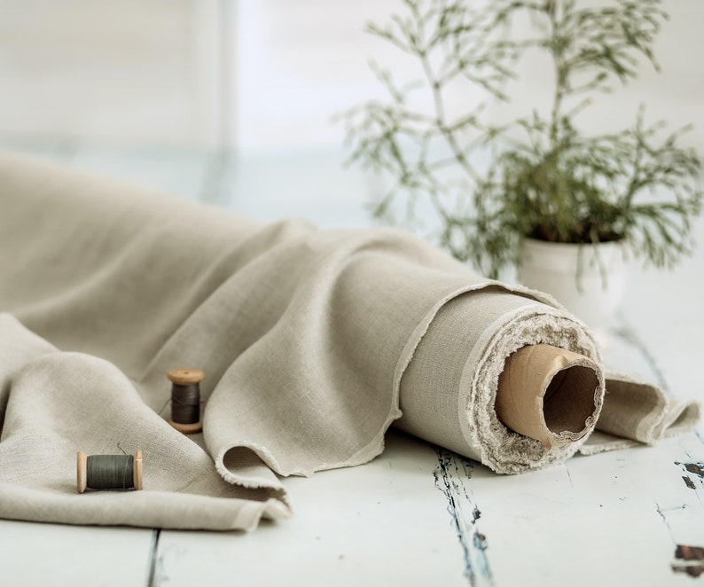 Puede incluir: Un rollo de tela de lino beige con un borde sin rematar, parcialmente desenrollado. Dos carretes de hilo de madera est&aacute;n en primer plano. Una peque&ntilde;a planta en maceta est&aacute; en el fondo, lo que sugiere un tema artesanal o textil.