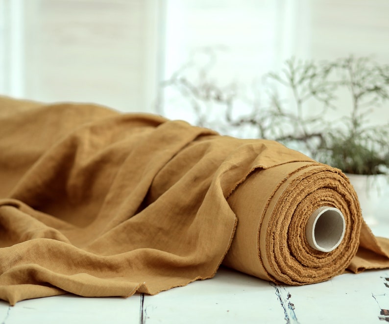 May include: A roll of tan-colored linen fabric. The fabric is loosely draped over a white surface, with a portion rolled up, revealing the texture and layers. The background includes a window and greenery.