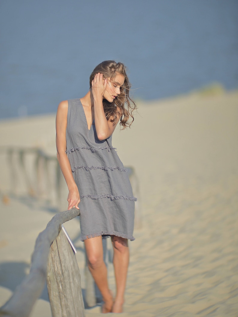 May include: A woman in a gray linen dress stands by a wooden fence on a sandy beach. The dress has a v-neck and a tiered skirt.