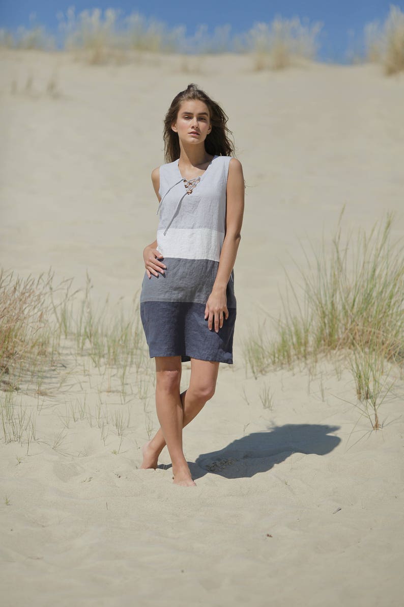 May include: A woman wearing a sleeveless, gray, white, and blue striped linen dress with a lace-up neckline, standing in a sandy desert setting.
