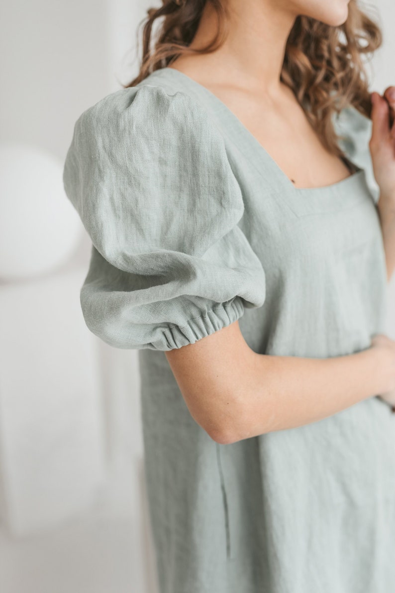May include: A sage green linen dress with a square neckline and puffed sleeves. The dress has a relaxed fit and is made of a lightweight fabric.
