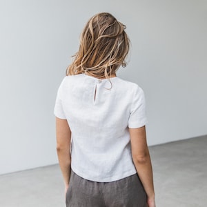 May include: A white linen top with a short sleeve and a button closure in the back. The top is cropped and has a relaxed fit.