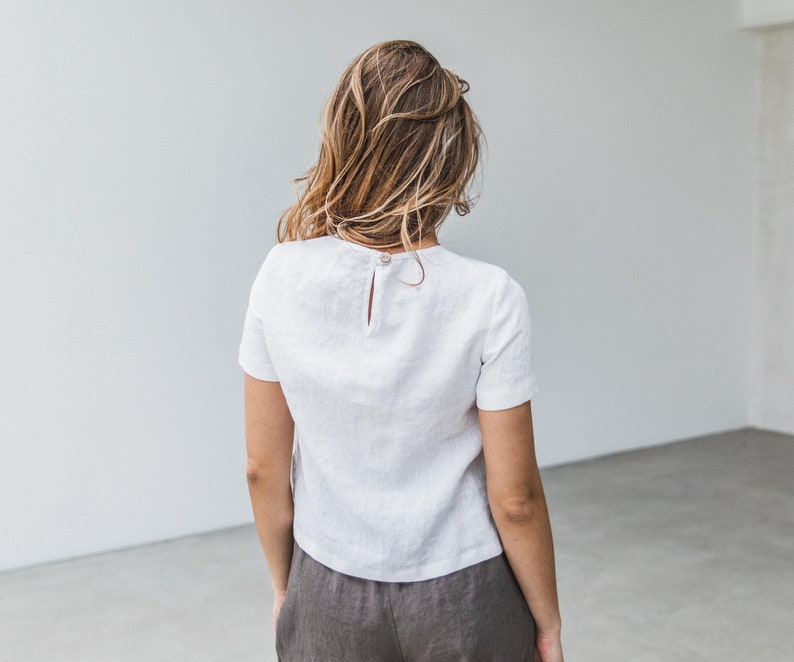 May include: A white linen top with a short sleeve and a button closure in the back. The top is cropped and has a relaxed fit.