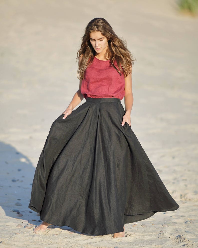 May include: A woman wearing a sleeveless, dark red top and a long, flowing black skirt. The skirt has a high waist and pockets. The outfit is made of a lightweight fabric, suitable for warm weather. The model is standing on a sandy beach.