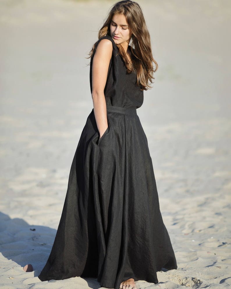 May include: A sleeveless black maxi dress with a wide skirt and pockets. The dress is made of a flowing fabric and is worn outdoors on a sandy beach. The model is wearing the dress.
