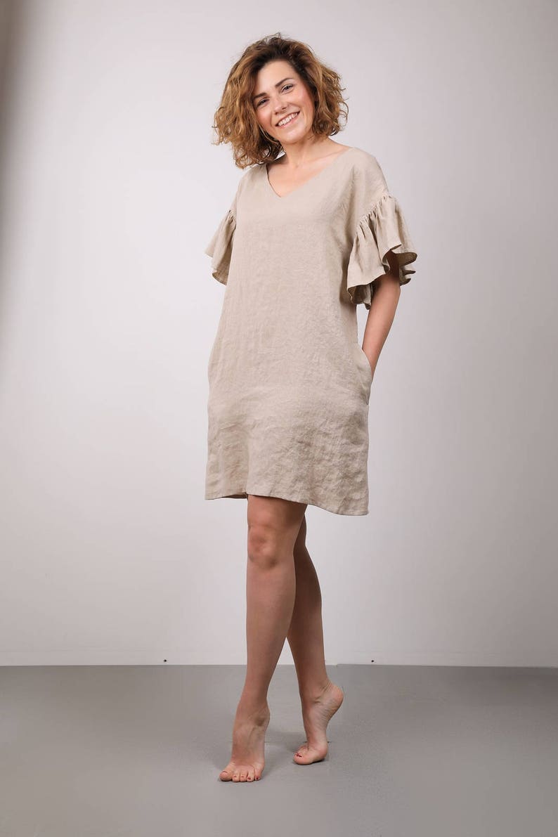 Op de afbeelding: Een vrouw draagt een beige linnen jurk met geruite mouwen. De jurk heeft een v-hals en zakken. Ze staat op een grijze vloer met een witte muur op de achtergrond.