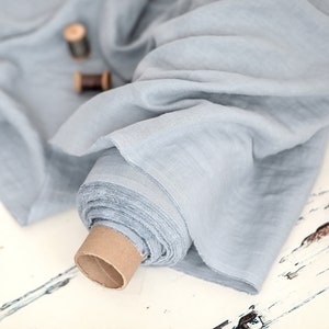 May include: A roll of light blue fabric, possibly linen or cotton, with a soft, flowing texture. The fabric is partially unrolled, revealing its layered edges. Two wooden spools of thread are visible in the background, suggesting sewing or crafting.