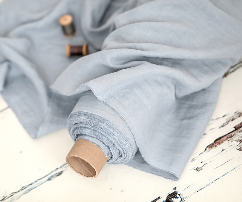 May include: A roll of light blue fabric, possibly linen or cotton, with a soft, flowing texture. The fabric is partially unrolled, revealing its layered edges. Two wooden spools of thread are visible in the background, suggesting sewing or crafting.