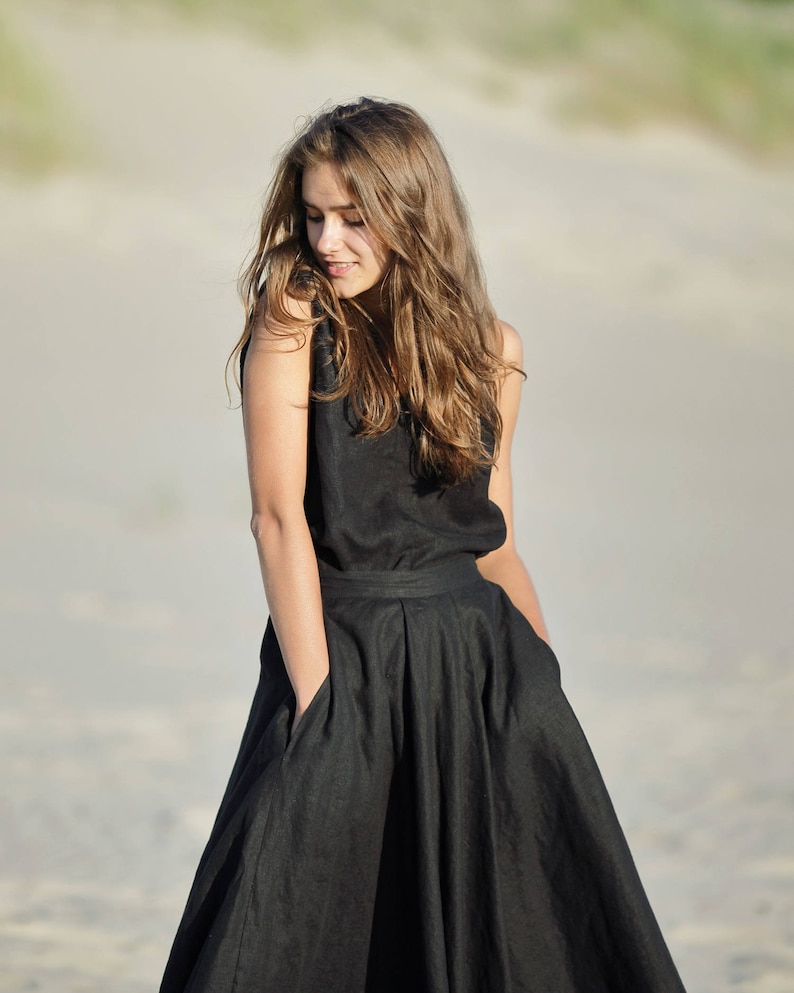 May include: A sleeveless black dress with a full skirt and pockets. The dress is made of a dark fabric and is worn outdoors. The model has long, light brown hair and is looking down.