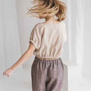 May include: A woman wearing a beige linen top with a gathered elastic waistband and brown striped linen pants. Her hair is blonde and flowing.