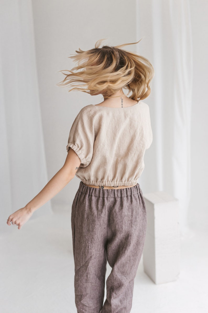 May include: A woman wearing a beige linen top with a gathered elastic waistband and brown striped linen pants. Her hair is blonde and flowing.