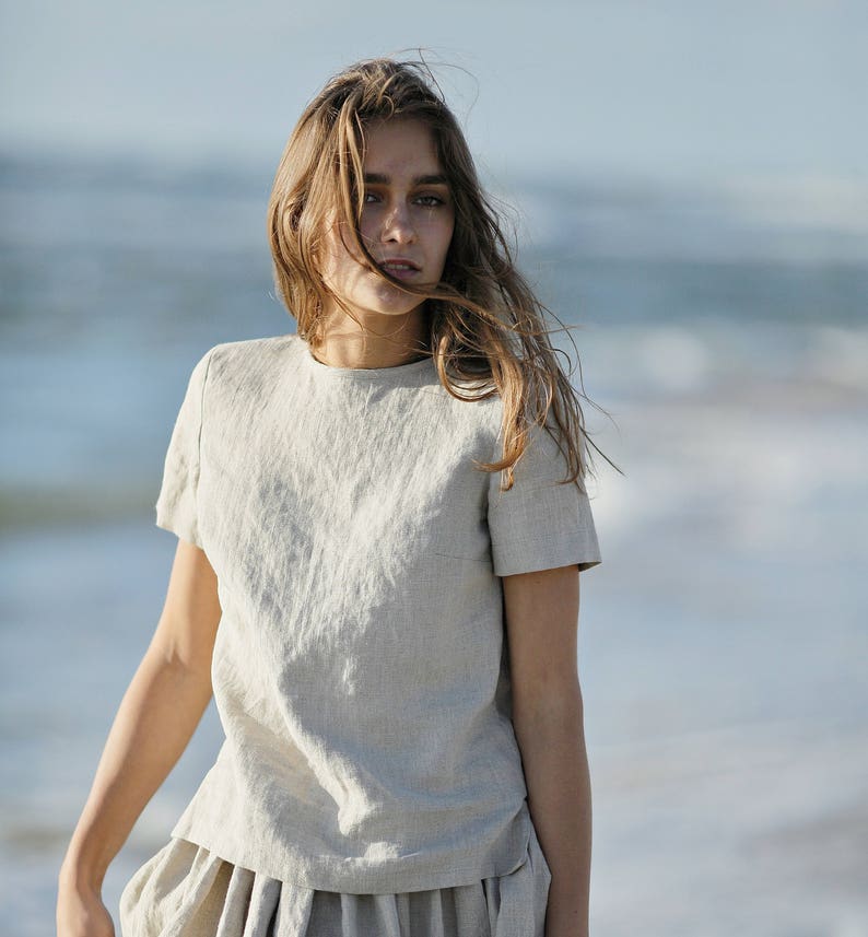 K&ouml;nnte beinhalten: Ein hellbeige Leinenoberteil mit kurzen &Auml;rmeln und ein passender Rock. Das Outfit besteht aus einem strukturierten Stoff und wird von einer Person mit langem, flie&szlig;endem Haar getragen. Der Hintergrund ist eine verschwommene Ansicht des Ozeans.