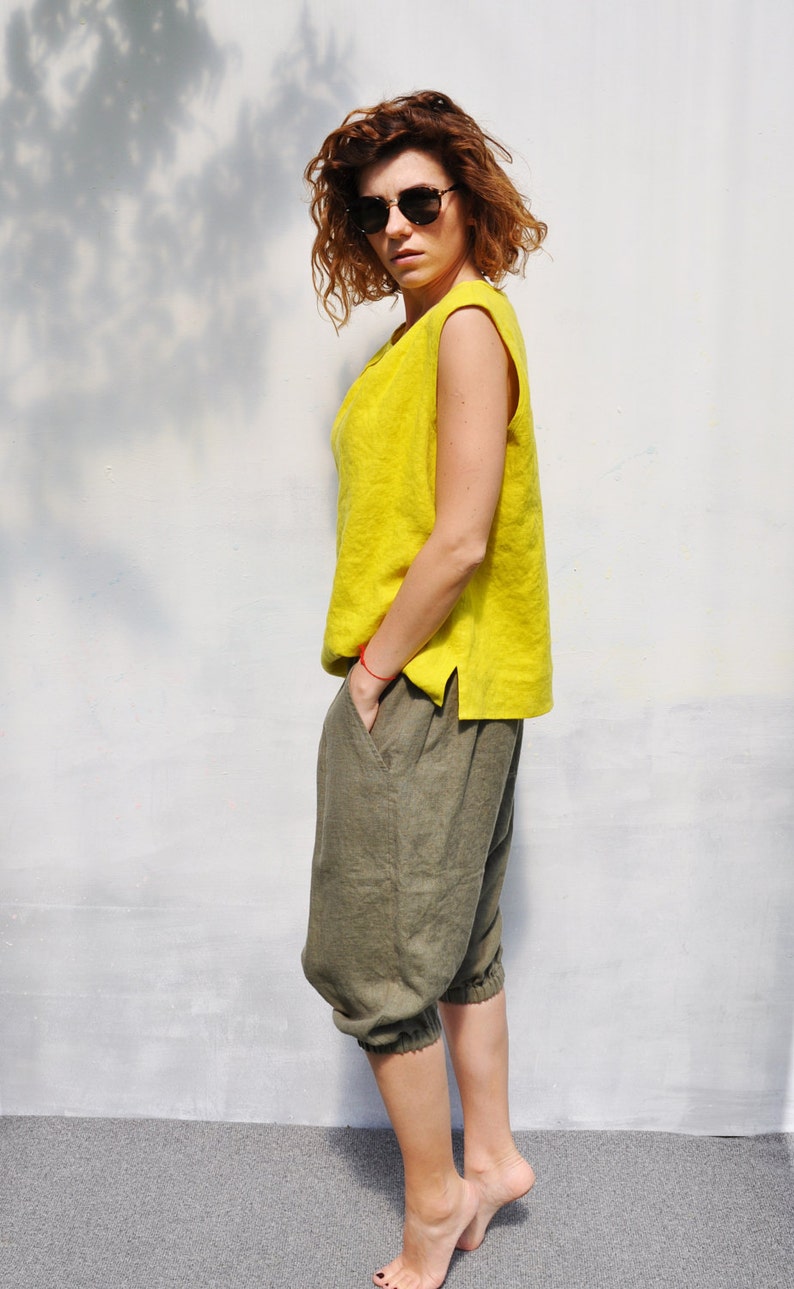 May include: A woman wearing a bright yellow sleeveless top and olive green linen trousers. She is standing in front of a white wall and has her hand in her pocket.