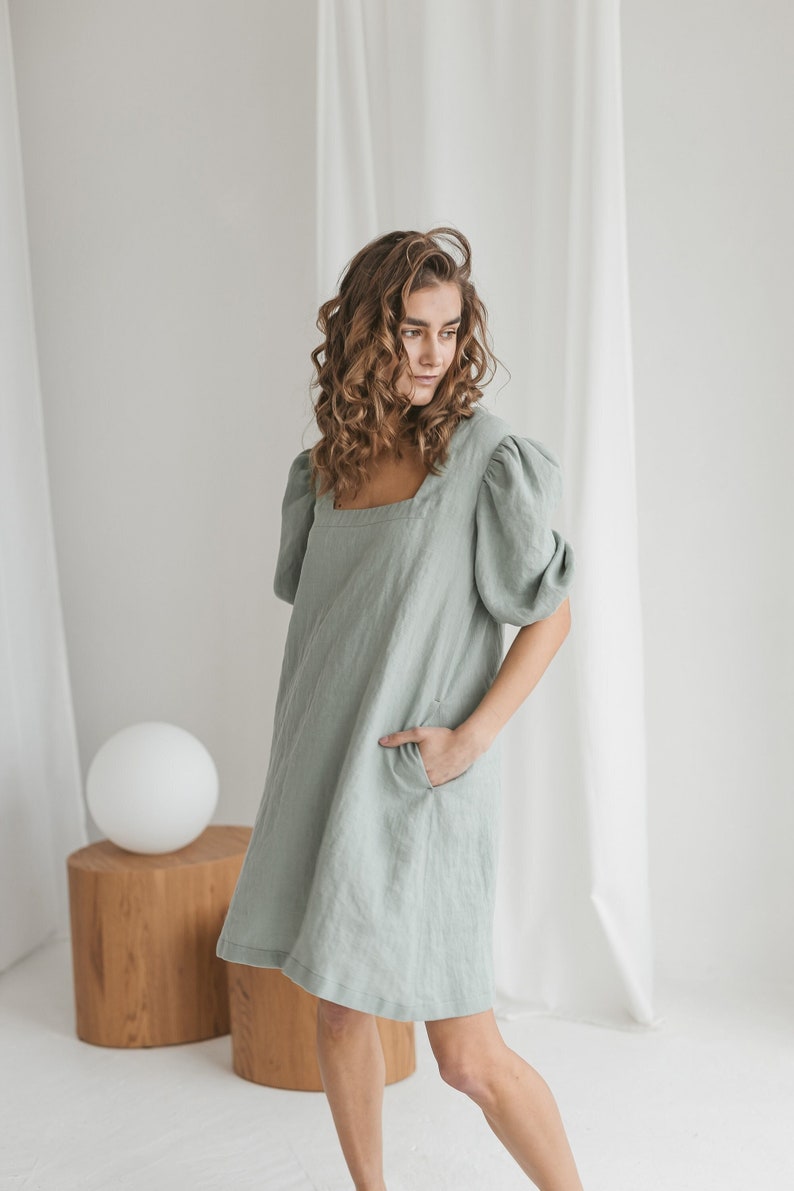 May include: A woman wearing a sage green linen dress with puffed sleeves and a square neckline. The dress has a relaxed fit and pockets on the sides.