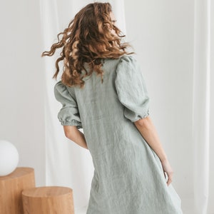 May include: A woman wearing a sage green linen dress with puffed sleeves. The dress has a relaxed fit and a slightly A-line silhouette.