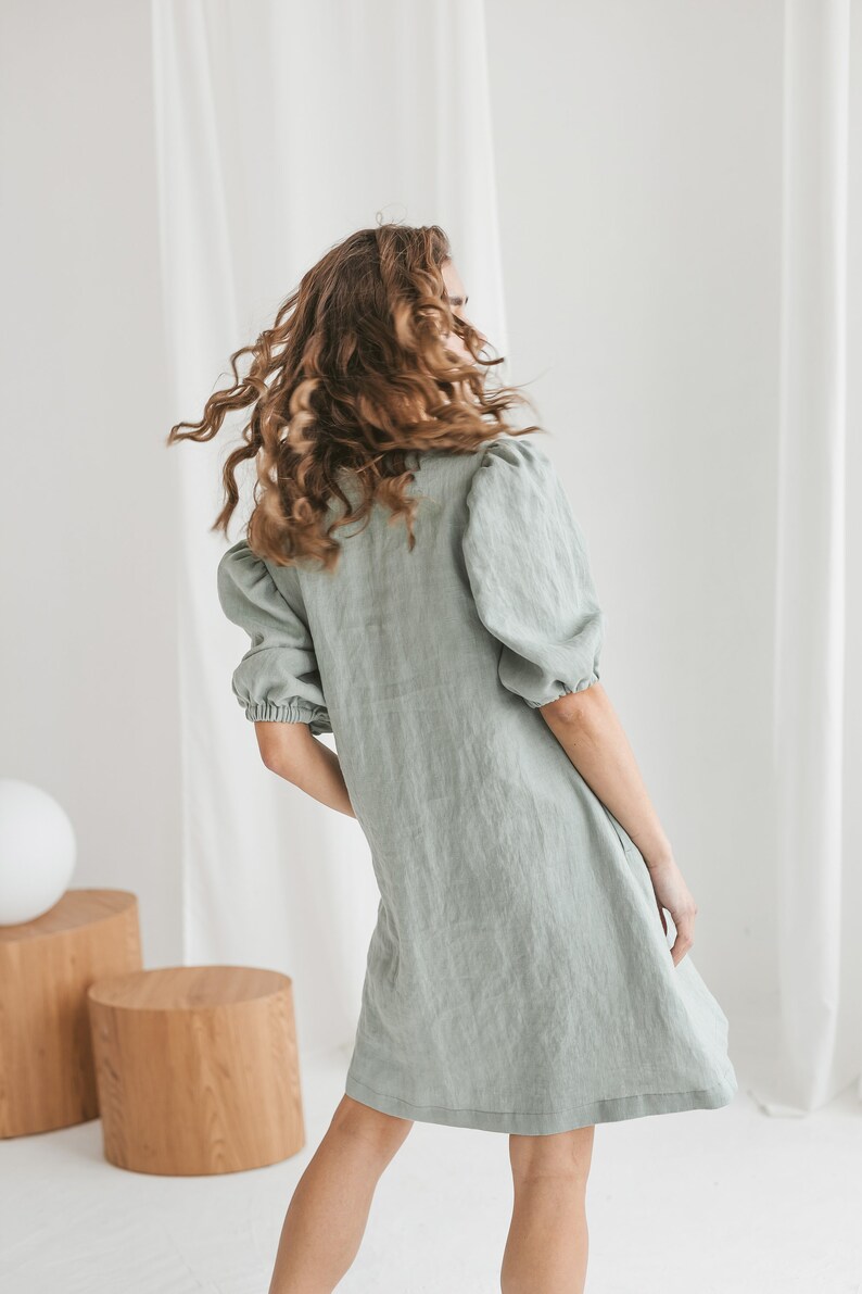 May include: A woman wearing a sage green linen dress with puffed sleeves. The dress has a relaxed fit and a slightly A-line silhouette.