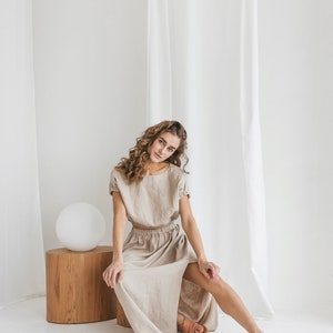 May include: A woman wearing a light brown linen dress with a slit in the skirt. She is sitting on a wooden stool in front of a white backdrop.