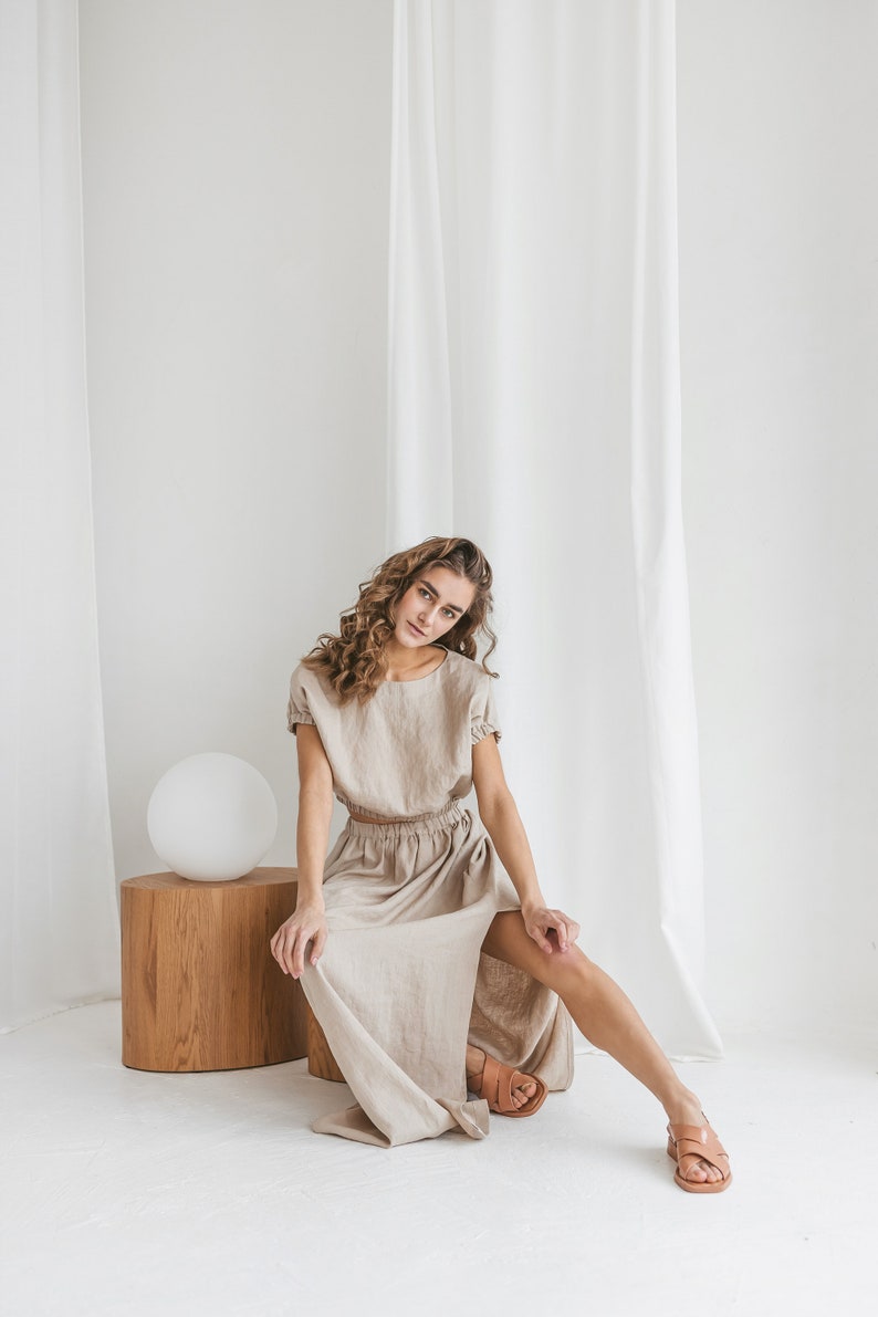 May include: A woman wearing a light brown linen dress with a slit in the skirt. She is sitting on a wooden stool in front of a white backdrop.