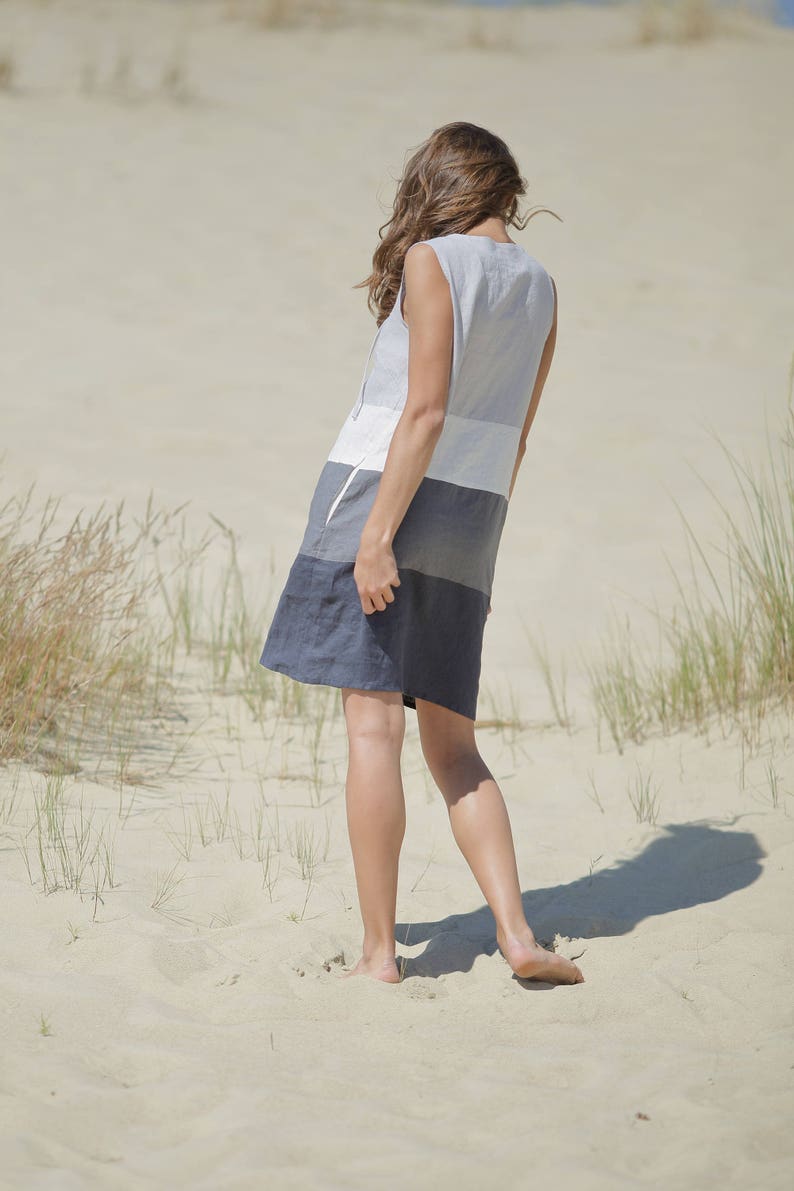 May include: A woman wearing a sleeveless dress with horizontal stripes in shades of gray and white. She is walking barefoot on a sandy beach.