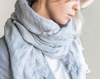 May include: A light blue linen scarf with frayed edges. The scarf is wrapped around a person's neck and is worn over a white shirt and a gray blazer.