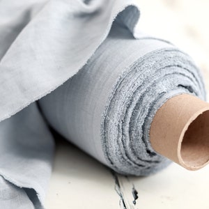 May include: A roll of light blue fabric, unraveled to show the texture and weave. The fabric is wrapped around a cardboard tube, with the edges of the fabric frayed. The color is a soft, muted blue, suitable for various sewing projects.