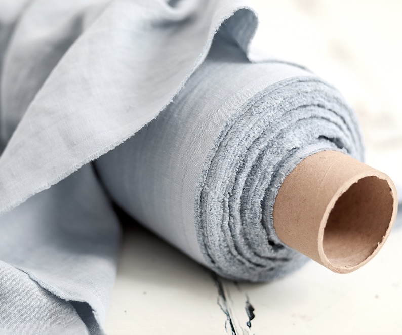 May include: A roll of light blue fabric, unraveled to show the texture and weave. The fabric is wrapped around a cardboard tube, with the edges of the fabric frayed. The color is a soft, muted blue, suitable for various sewing projects.