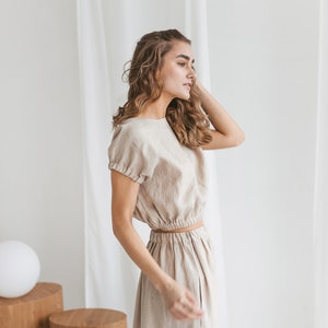 May include: A woman wearing a light beige linen dress with a fitted bodice and a flowing skirt. The dress has a cinched waist and short sleeves.