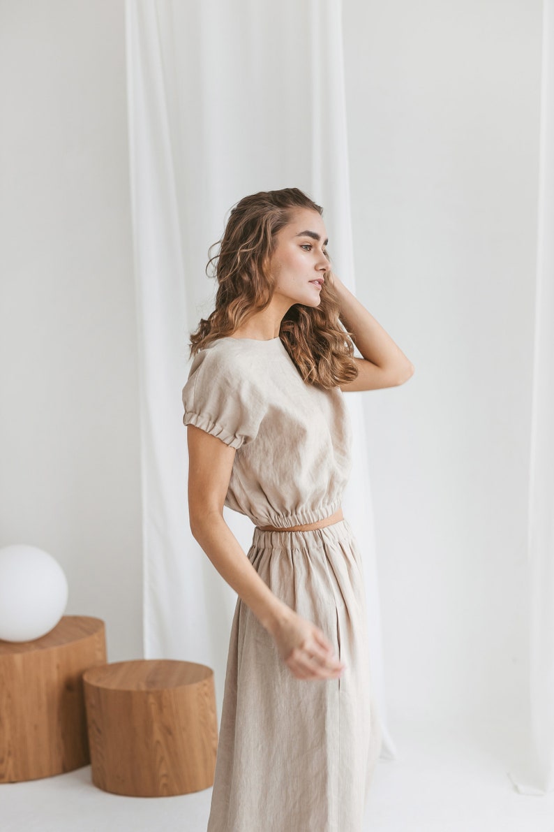 May include: A woman wearing a light beige linen dress with a fitted bodice and a flowing skirt. The dress has a cinched waist and short sleeves.