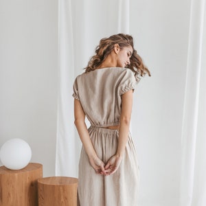 May include: A woman wearing a light brown linen dress with a fitted bodice and a flowing skirt. The dress has a scoop neckline and short sleeves.