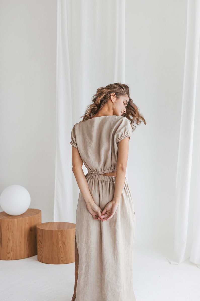 May include: A woman wearing a light brown linen dress with a fitted bodice and a flowing skirt. The dress has a scoop neckline and short sleeves.