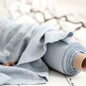 May include: A roll of light blue linen fabric. The fabric is loosely rolled around a cardboard core, with a portion of the fabric draped over the roll. Two wooden spools are in the foreground. The fabric has a soft, textured appearance.