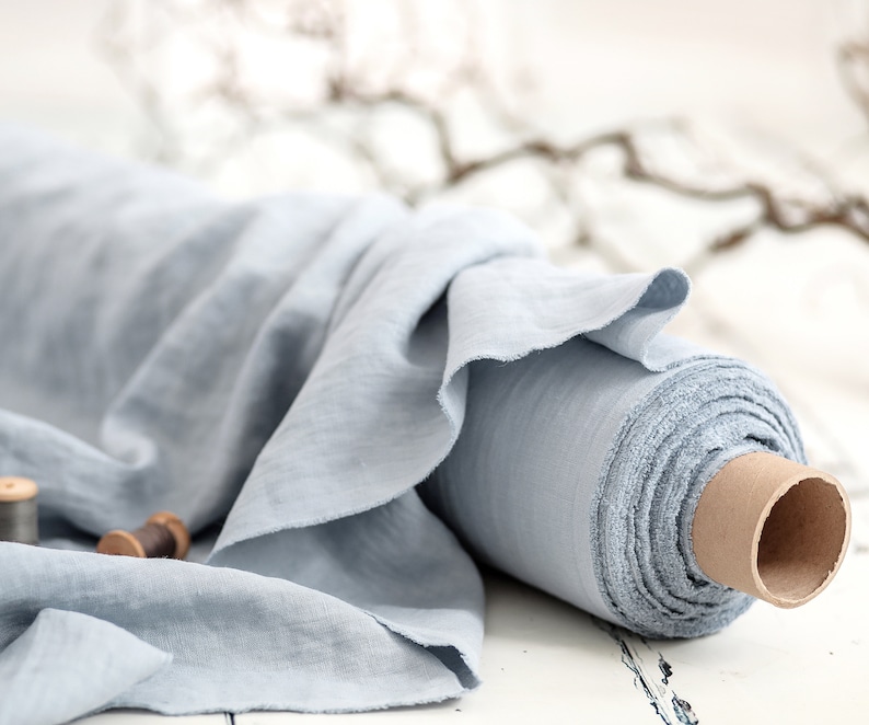 May include: A roll of light blue linen fabric. The fabric is loosely rolled around a cardboard core, with a portion of the fabric draped over the roll. Two wooden spools are in the foreground. The fabric has a soft, textured appearance.