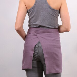 May include: A purple linen apron with a back slit and a tie closure. The apron is worn over a gray tank top and gray jeans.