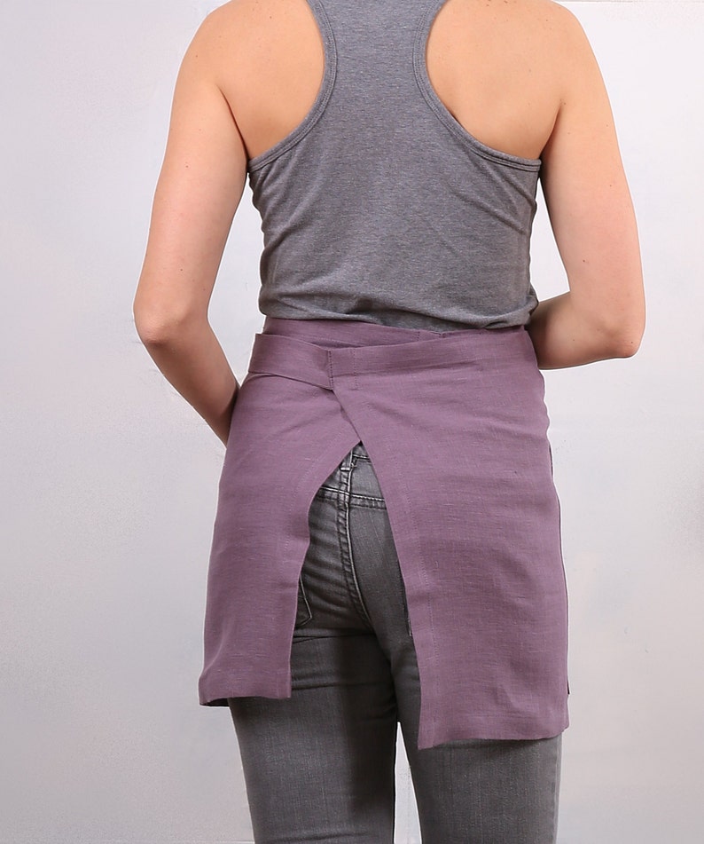 May include: A purple linen apron with a back slit and a tie closure. The apron is worn over a gray tank top and gray jeans.