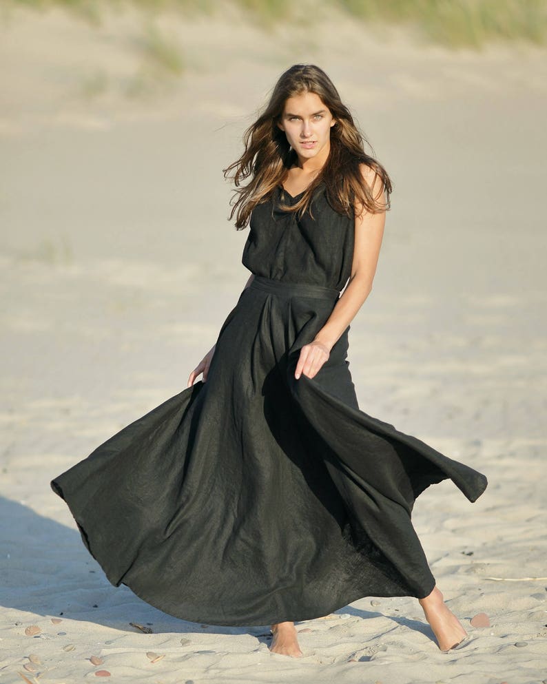 May include: A black linen dress with a V-neck top and a flowing skirt. The dress is worn on a sandy beach. The model is barefoot and the dress is billowing in the wind.