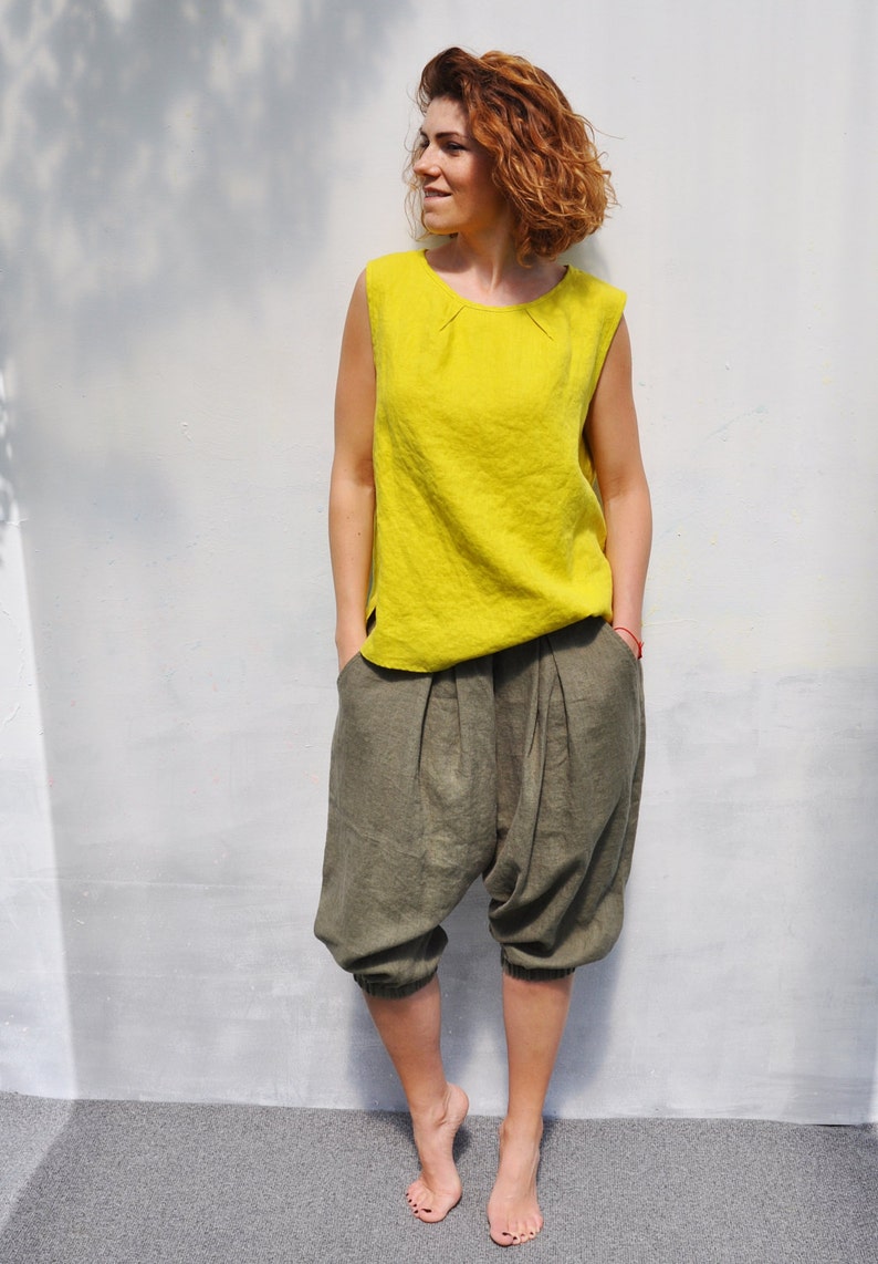 May include: A woman wearing a bright yellow sleeveless top and olive green linen trousers. She is standing in front of a white wall.